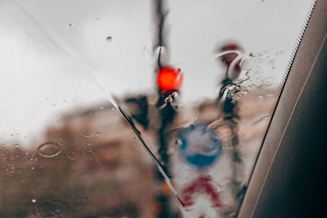 Blurred red traffic light seen through raindrops on a car windshield in a city setting.