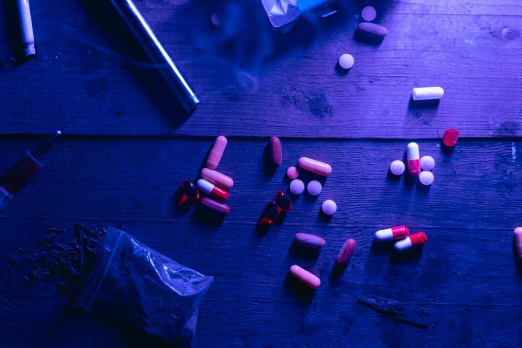 pexels-photo-7230391-7230391 A flat lay of various drugs including pills, capsules, and a syringe on a wooden surface under moody blue lighting.